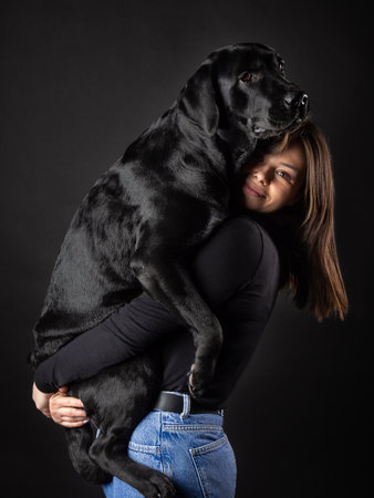 A girl holds a Labrador Retriever dog in her arms. Taken close-up in a photo studio, on a black background.の写真素材