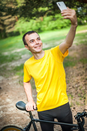 A young man taking a selfie with his Bicycle. In a public park, among trees and vegetation.の写真素材