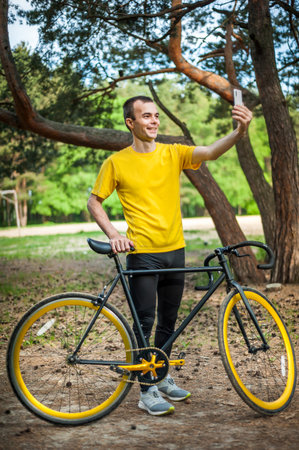 A young man taking a selfie with his Bicycle. In a public park, among trees and vegetation.