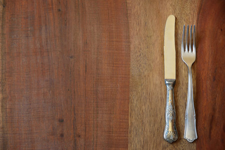 Background from a table top of a rustic wooden table on which lie an old decorated fork and an old decorated knifeの写真素材