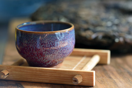 Closeup of a blue speckled ceramic teacup, on a wooden board, with a pressed cake of tea leaves in...