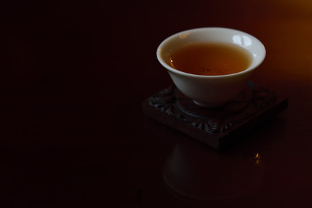 A small white Chinese teacup with black fresh tea in it stands against brown and dark background and is reflected in golden light on a tabletop with space for textの写真素材