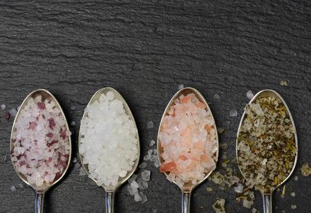 Close-up of four silver teaspoons lying on a black slate plate and in which various salt mixtures and salt crystals lie in the kitchenの写真素材