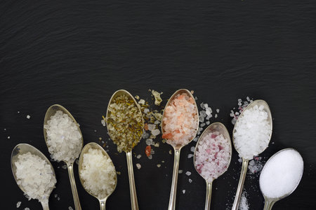 Close-up of eight silver teaspoons lying on a black slate plate and in which various salt mixtures and salt crystals lie in the kitchen with space for textの写真素材