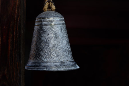 A metal bell hangs rustic on a cord next to a wooden beam against a dark background with space for textの写真素材