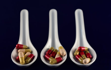 Three white large spoons for medicine, filled with various colorful capsules and medicines, side by...