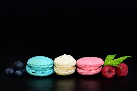 Three sweet macarons, in the colors of the French flag, lie side by side against a dark background, with blueberries and raspberriesの写真素材