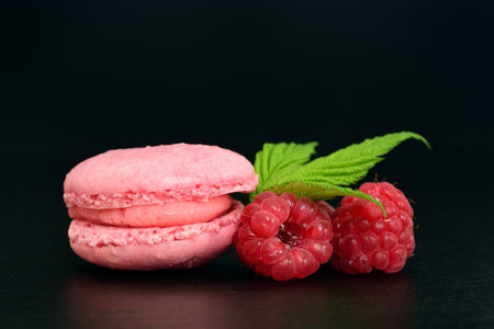 A pink macaron with raspberries and green leaves against a dark backgroundの写真素材
