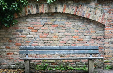 An old wooden bench stands in front of a historic wall made of burnt red bricks with a round arch and ivyの写真素材