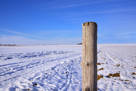 A weathered fence post stands in a wide winter landscape in Bavaria, against snow and a blue skyの写真素材