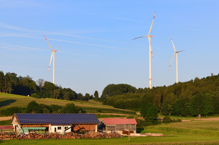 On August 25, 2021, the evening sun shines on a farm near Augsburg, Germany, on whose barn there is a solar system. In the background a lot of nature, sky and three large wind turbinesのeditorial素材
