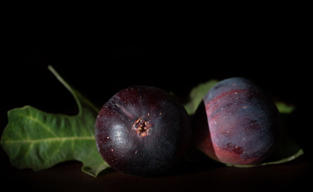 Dark shot of two red figs lying side by side on a table. A few fig leaves in the background. The room is dark.の写真素材