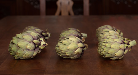 Several artichokes lie next to each other on a rustic wooden table. In the background an old wooden cupboard and a detail of a wooden chair.の写真素材