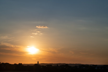 The sun sets over a small village in Bavaria. You can see the silhouette and the church tower of the village. It is evening. The sun shines red, the sky is dark blueの写真素材