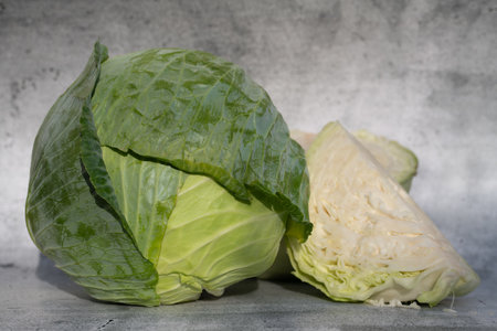 Fresh white cabbage lies against a gray background. A head of cabbage is still whole, with green leaves, in the foreground. Next to it lies a piece of cut cabbage with light structures.の写真素材