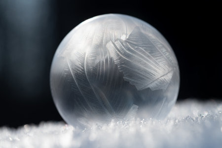 Close-up of a shimmering frozen soap bubble lying on snow crystals. The background is dark. The light shines from the side.の写真素材
