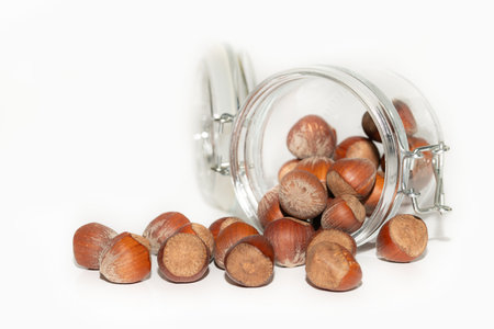 Close-up of hazelnuts that have spilled out of an overturned jar. The nuts and the storage jar are cropped against a light-colored background.の写真素材