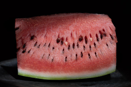 Close-up of a fresh slice of watermelon lying on a metal plate. The background is dark. The seeds are clearly visible in the red flesh.の写真素材