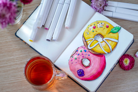 sketching donuts in a notebook. All placed on a wooden table with a transparent cup of tea, notebook, markers, and flowers. Top viewの写真素材