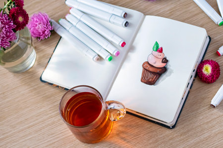 Sketching cupcake in a notebook. All placed on a wooden table with a transparent cup of tea,...