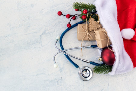Santa Claus hat and stethoscope, Christmas gifts, and a branch of Christmas tree with red Christmas ball on a white concrete table. Xmas. Copy space. Flat layの写真素材