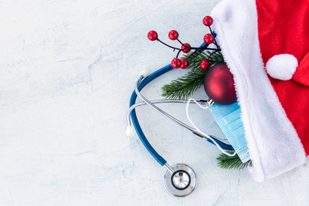Santa Claus hat and stethoscope, blue protective mask and branch of Christmas tree with red Christmas ball on a white concrete table. Copy space. Flat layの写真素材