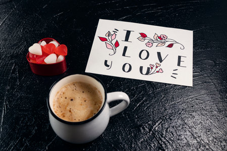 Mug of coffee, red and white heart-shaped marmalade and a valentine card with the word I love you on black background. Valentine's Day breakfast. Valentine's day concept. Soft focusの写真素材