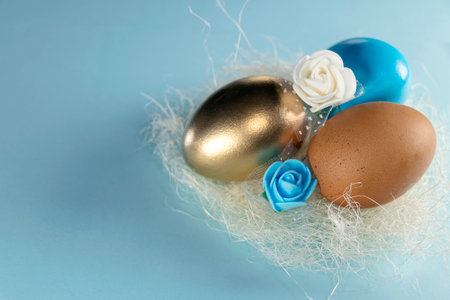 Gold, blue and brown eggs in the nest. Tree aggs in nest. Happy Easter concept. Painted eggs in a nest with a blue and white rose on a pastel blue background. copy spaceの写真素材