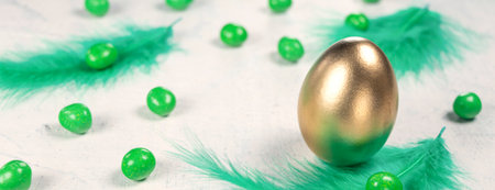Banner with Golden egg on a white concrete background with green feathers and green chocolates in the shape of an egg. Happy Easter concept. Soft focusの写真素材