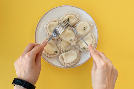 female hands with a knife and a fork eat delicious, fresh, homemade italian ravioli on a clay plate on a yellow background. Top viewの写真素材