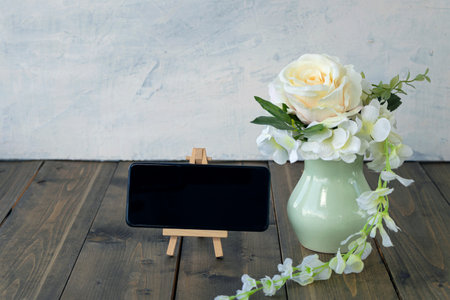 Bouquet from peach rose and white hydrangea in pastel green vase near empty mobile phone on wooden...