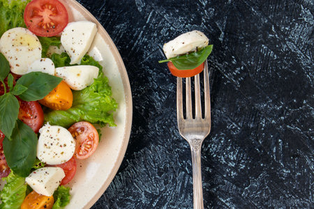 Caprese salad with tomatoes, mozzarella, basil and olive oil on a black texture background. fork and...
