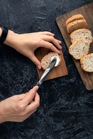 female hands smear cream cheese on bread with a knife on a black texture background. the process of...