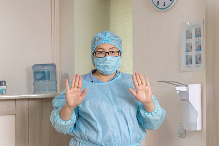 A medical worker in a disposable medical suit, a medical cap and a mask with glasses, shows the treated hands with a disinfectant solution next to the elbow dispenser. soft focusの写真素材