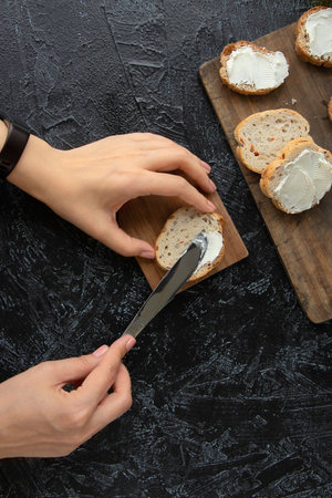 the process of making sandwiches or snacks. female hands smear cream cheese on bread with a knife on a black texture background. Soft focus. top view . flat layの写真素材