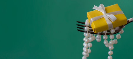 banner with decorations on a fork, white pearl beads and a yellow gift on a green background. festive food concept for christmas, new year, anniversary or wedding. copy spaceの写真素材