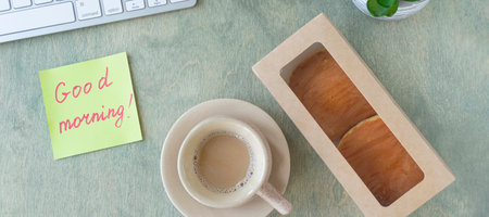 banner with flat lay with of the desktop. note with the words GOOD MORNING, pancakes in paper box, cup of coffee, keyboard, cactus and mause on a wooden table. concept of good start to day. top viewの写真素材