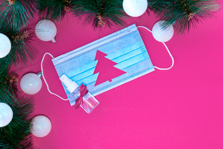 Blue medical protective mask with cut-out silhouette of Christmas tree and sanitizer with scarlet bow on scarlet background. Christmas tree branches near mask. Christmas and Happy New Year conceptの写真素材