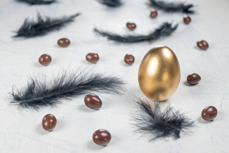 golden egg on a white concrete background with black feathers and chocolates in the shape of an egg. Happy Easter concept. Soft focusの写真素材