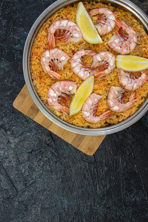 Vertical photo with a top view of a frying pan with paella on a wooden board on a black textured background. flat lay Classic dish of Spain, seafood paella, copy space. soft focus.の写真素材