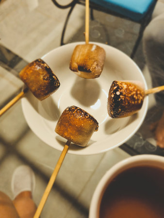Vertical photo of golden-brown toasted marshmallows on wooden skewers with soft focus, capturing a cozy and warm outdoor snack atmosphere.の写真素材