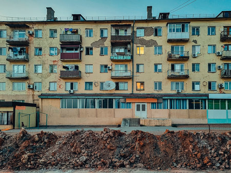 Photo of a worn Soviet-era five-story residential building with peeling plaster, lit windows, hanging laundry, and dug-up ground in front.の写真素材