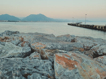 Peaceful coastal landscape with large rocks in the foreground, calm sea, distant mountains and an empty wooden pier stretching into the water. Bright natural daylight. Soft focusの写真素材