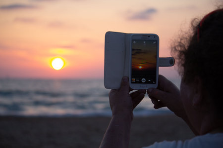 Photographing with smartphone in hand. Travel concept. Sunset on the sandy beach. Vacation at seaの写真素材