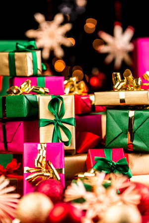 Pile of plain Christmas presents between blurred baubles and straw stars. Vibrant colors. Magenta, gold, green and red. Tightly framed. Narrow depth of field. Bokeh over pitch black background.の写真素材