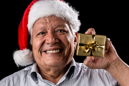 Aged, happy man is cracking a smile while holding up a small wrapped golden present in his left hand. He is wearing a red and white Father Christmas cap. Focus on eyes. Black background. Ample DOF.の写真素材