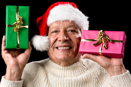 Friendly old man with red Santa Claus hat and warm pullover. He is raising a green and a magenta wrapped present. Each gift decorated with golden bow. Isolated on black. Gift-giving theme. Ample DOF.の写真素材
