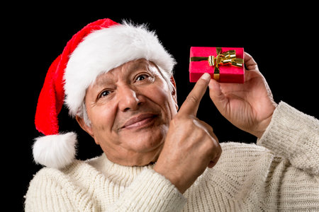 Pensive aged man in warm pullover and red Santa Claus cap. He is pointing his right index finger at a small, red, wrapped gift held on eye-level in his left hand. Isolated on black. Gift-giving theme.の写真素材