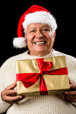 Cheerful aged gentleman offering a golden wrapped gift, which he is holding in front of his chest with both hands. He is wearing a red Santa Claus cap and a warm, white pullover. Isolated on black.の写真素材