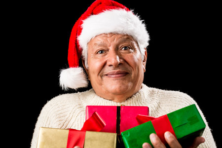 Friendly, but perplexed looking male pensioner with Santa Claus cap. He is carrying Christmas presents in front of his chest. Wrapped in green, red and gold. Isolated on black. Gift giving theme.の写真素材